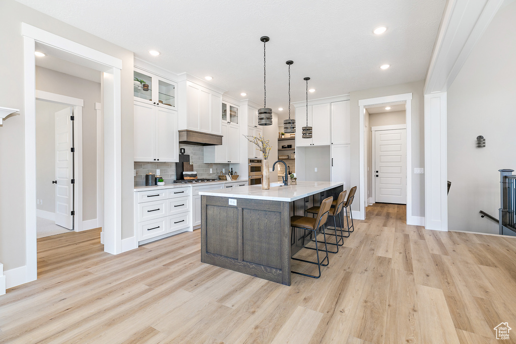Kitchen featuring light wood-type flooring, custom exhaust hood, backsplash, white cabinets, and an island with sink