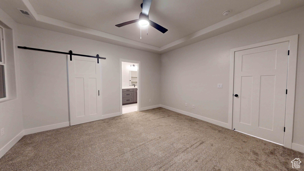 Unfurnished bedroom featuring light colored carpet, a barn door, a tray ceiling, and ceiling fan