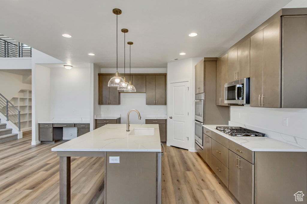 Kitchen with light hardwood / wood-style floors, appliances with stainless steel finishes, hanging light fixtures, a kitchen island with sink, and sink