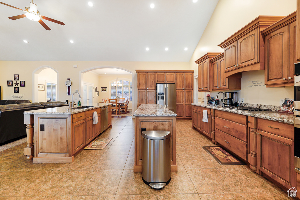Kitchen featuring lofted ceiling, ceiling fan with notable chandelier, light tile flooring, appliances with stainless steel finishes, and an island with sink