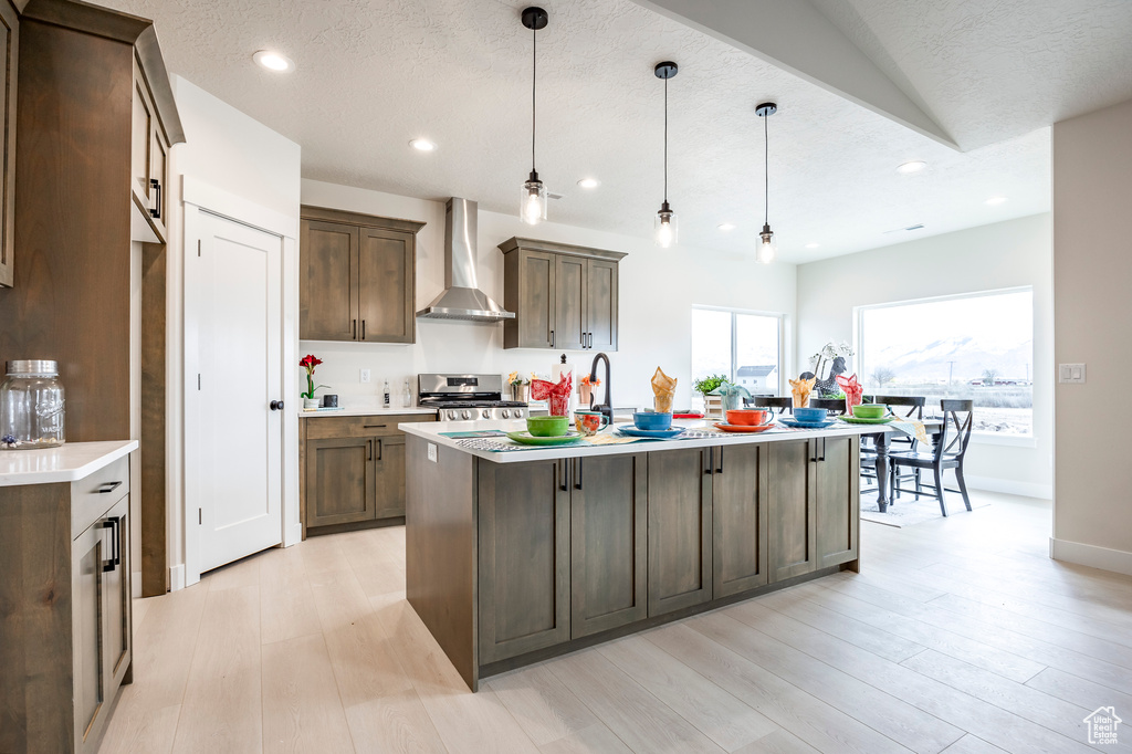 Kitchen featuring wall chimney exhaust hood, light hardwood / wood-style flooring, range, decorative light fixtures, and a center island with sink