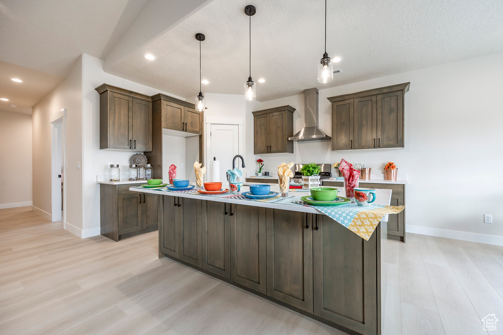 Kitchen with light hardwood / wood-style flooring, pendant lighting, wall chimney range hood, and a kitchen island with sink