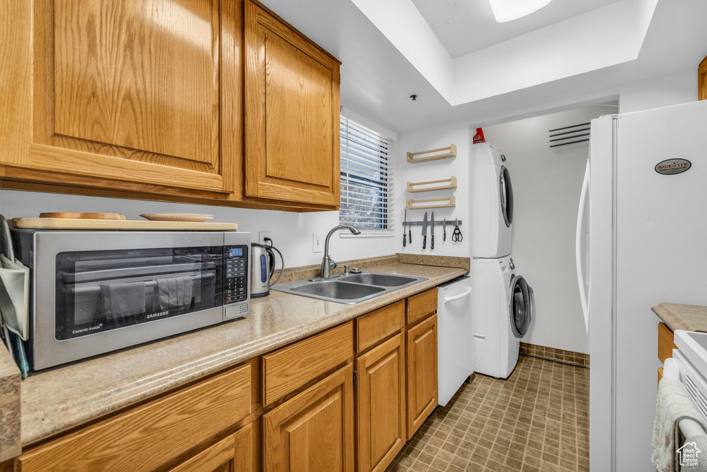 Kitchen featuring light tile flooring, white appliances, stacked washer and dryer, sink, and a tray ceiling