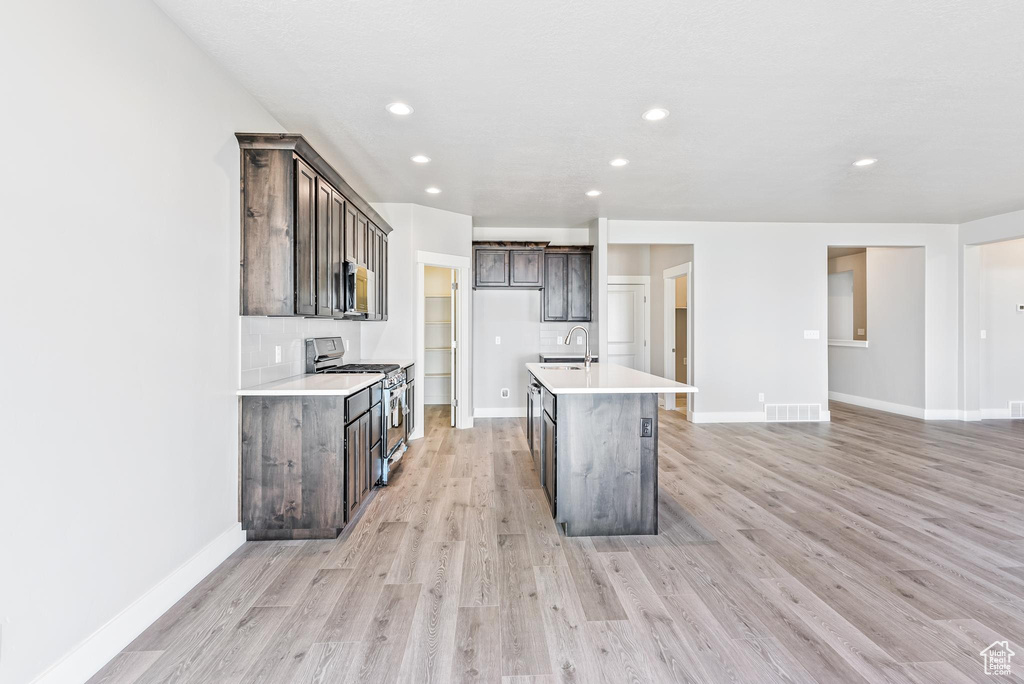 Kitchen featuring dark brown cabinetry, light hardwood / wood-style floors, tasteful backsplash, a kitchen island with sink, and appliances with stainless steel finishes