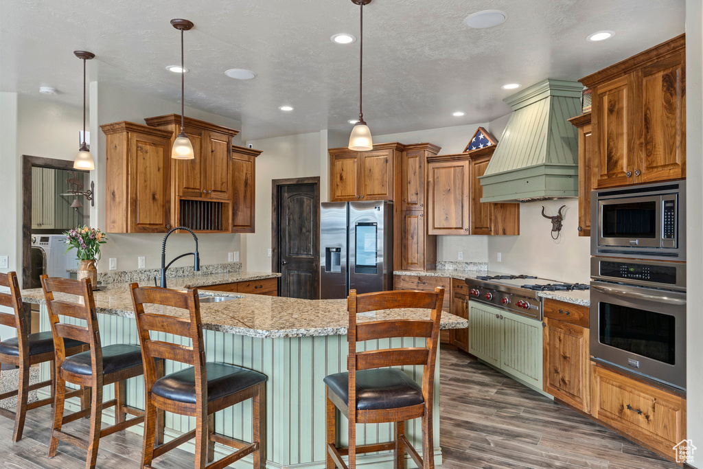 Kitchen with a breakfast bar area, appliances with stainless steel finishes, custom range hood, light stone counters, and dark hardwood / wood-style floors