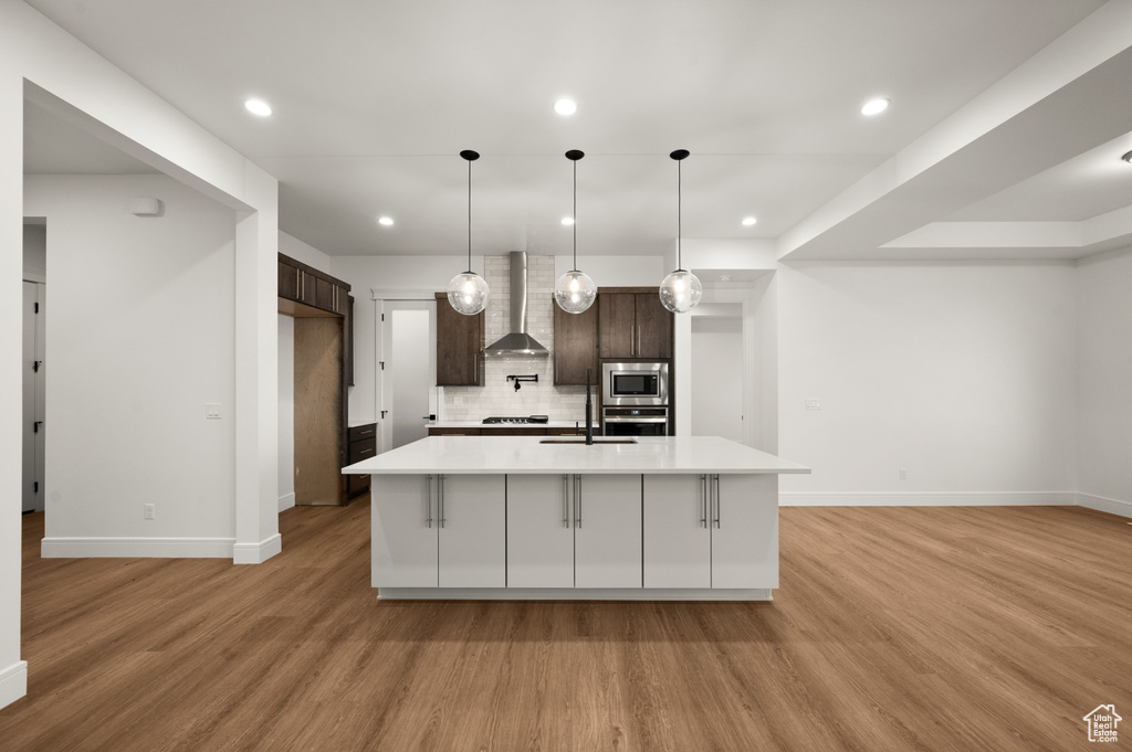 Kitchen featuring wall chimney range hood, light hardwood / wood-style floors, appliances with stainless steel finishes, tasteful backsplash, and dark brown cabinets