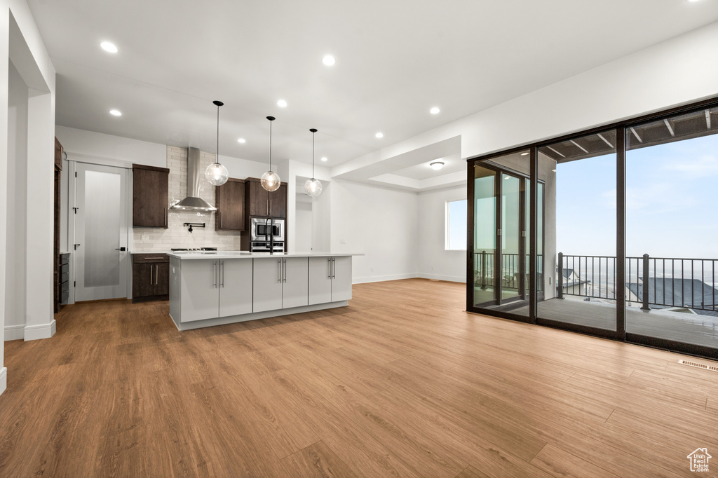 Kitchen with tasteful backsplash, dark brown cabinets, wall chimney range hood, and light wood-type flooring