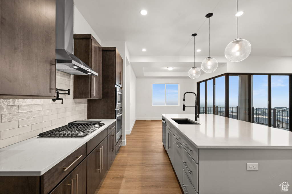 Kitchen featuring light hardwood / wood-style floors, wall chimney exhaust hood, backsplash, sink, and an island with sink