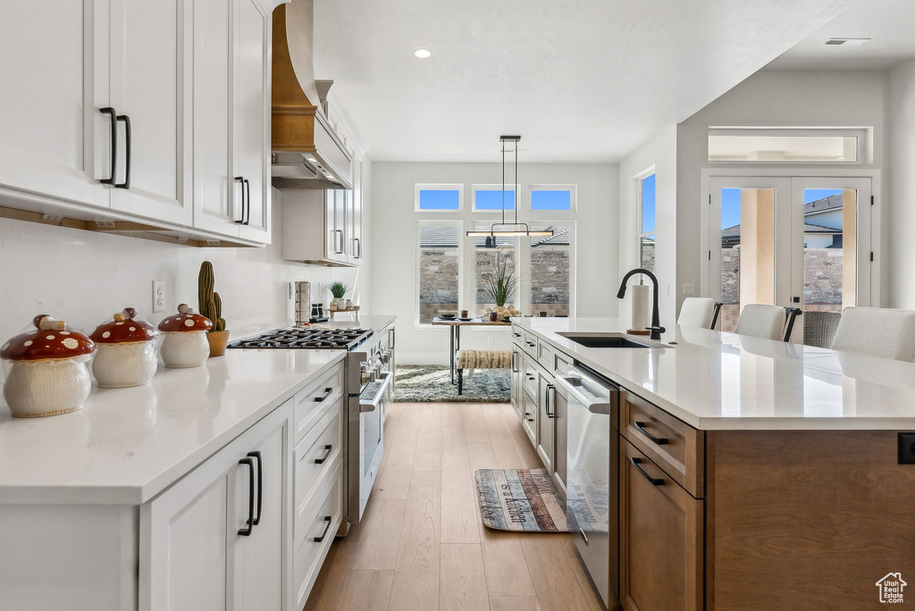 Kitchen featuring a sink, an island with sink, light wood-style flooring, stainless steel appliances, and custom range hood