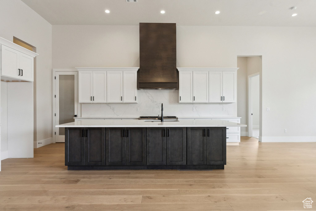 Kitchen with custom exhaust hood, light wood-style floors, a center island with sink, light countertops, and white cabinetry
