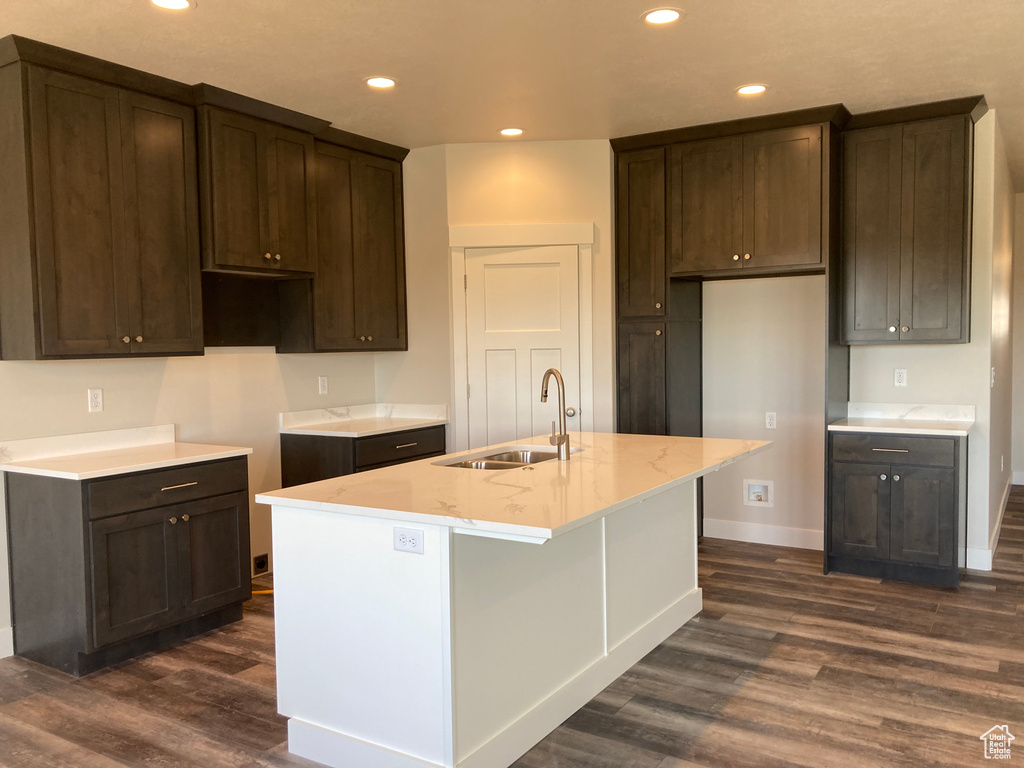 Kitchen with a center island with sink, sink, dark brown cabinetry, dark hardwood / wood-style flooring, and light stone countertops