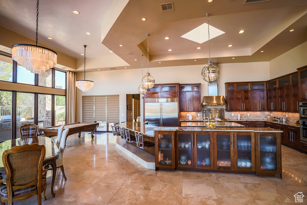 Kitchen featuring light stone counters, backsplash, a center island with sink, wall chimney range hood, and stainless steel appliances
