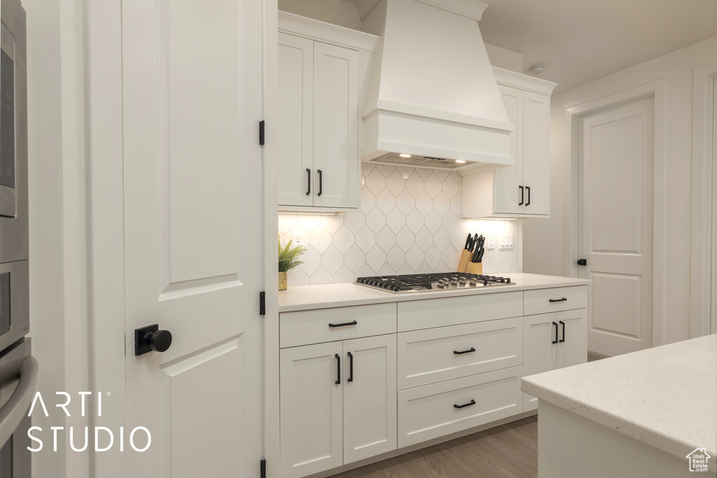 Kitchen with light wood-type flooring, white cabinetry, custom range hood, stainless steel gas stovetop, and backsplash