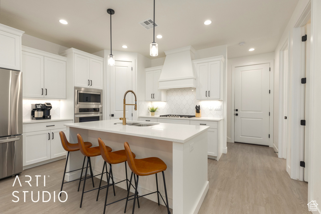 Kitchen with premium range hood, tasteful backsplash, appliances with stainless steel finishes, light hardwood / wood-style floors, and an island with sink