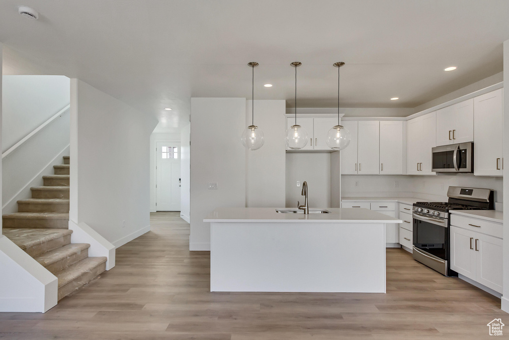 Kitchen with light wood-type flooring, white cabinets, hanging light fixtures, appliances with stainless steel finishes, and a kitchen island with sink