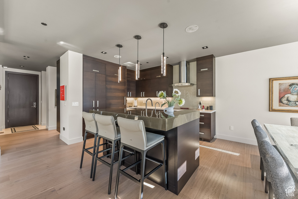 Kitchen featuring light wood-type flooring, tasteful backsplash, a breakfast bar, dark brown cabinetry, and wall chimney exhaust hood
