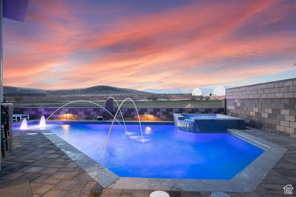 Pool at dusk featuring a mountain view, pool water feature, and an in ground hot tub
