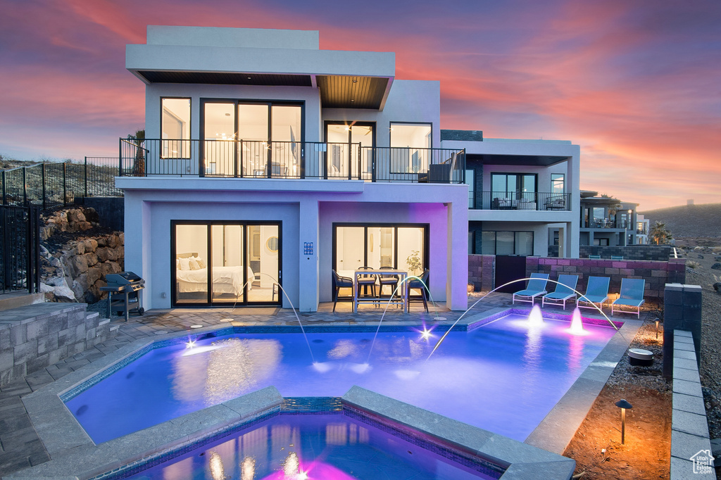 Back house at dusk featuring a pool with hot tub, a balcony, and pool water feature