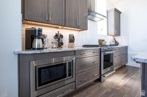 Kitchen featuring light stone counters, backsplash, dark hardwood / wood-style flooring, appliances with stainless steel finishes, and wall chimney exhaust hood