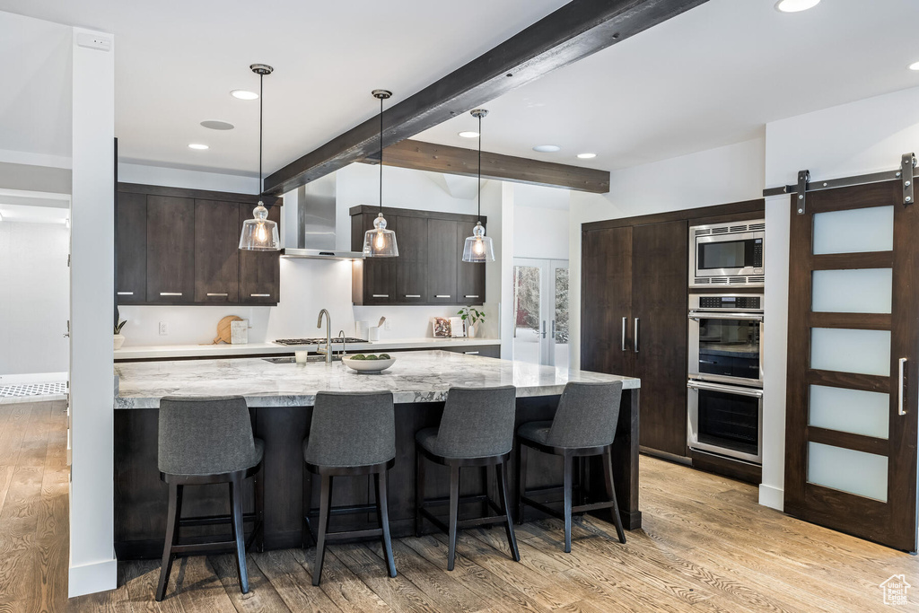 Kitchen with dark brown cabinets, a barn door, hanging light fixtures, light hardwood / wood-style flooring, and wall chimney range hood