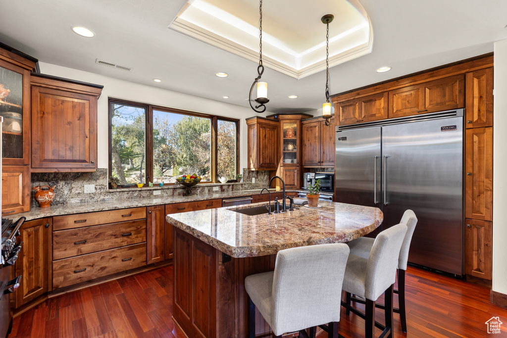 Kitchen featuring dark hardwood / wood-style floors, light stone countertops, stainless steel built in fridge, and a kitchen island with sink