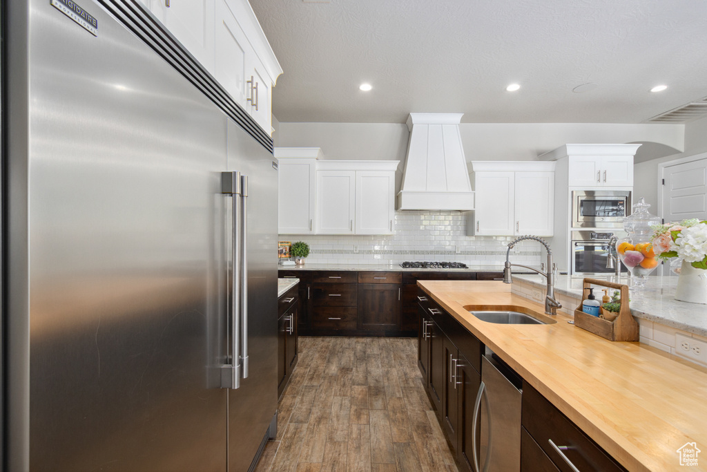 Kitchen with built in appliances, premium range hood, dark hardwood / wood-style flooring, tasteful backsplash, and sink