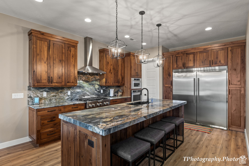Kitchen with light hardwood / wood-style floors, a center island with sink, tasteful backsplash, stainless steel appliances, and wall chimney range hood