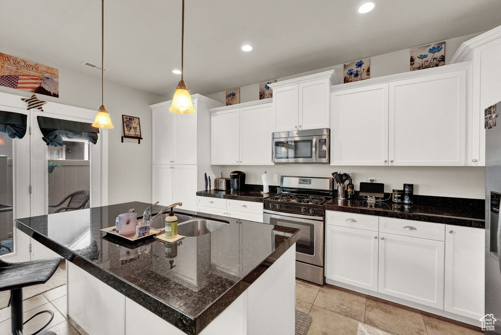 Kitchen featuring a kitchen island with sink, white cabinets, light tile floors, stainless steel appliances, and decorative light fixtures