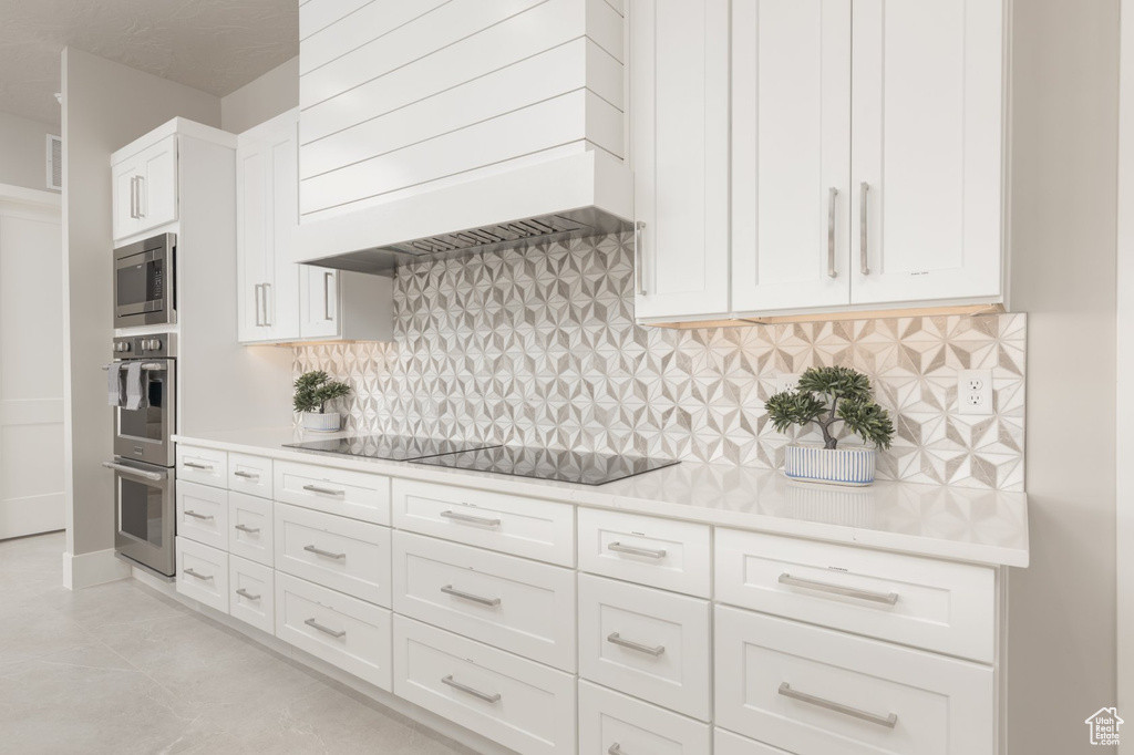 Kitchen featuring custom exhaust hood, stainless steel appliances, white cabinetry, backsplash, and light tile flooring