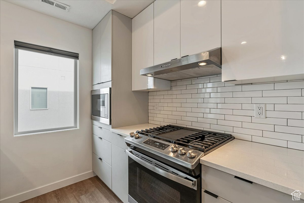 Kitchen with wall chimney exhaust hood, white cabinets, backsplash, appliances with stainless steel finishes, and light hardwood / wood-style flooring