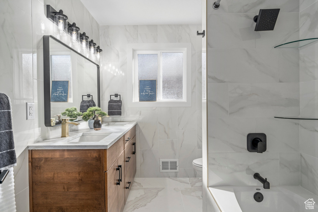 Full bathroom featuring tiled shower / bath combo, tile walls, toilet, dual bowl vanity, and tile flooring