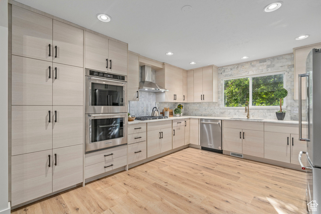 Kitchen with light wood-type flooring, backsplash, appliances with stainless steel finishes, wall chimney range hood, and sink