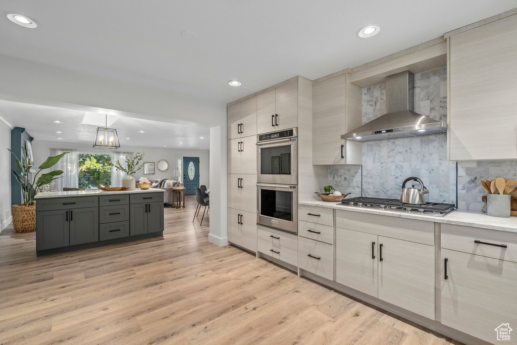 Kitchen featuring gray cabinets, light wood-type flooring, tasteful backsplash, stainless steel appliances, and wall chimney exhaust hood