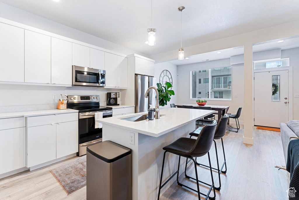 Kitchen featuring stainless steel appliances, white cabinets, hanging light fixtures, light hardwood / wood-style flooring, and an island with sink