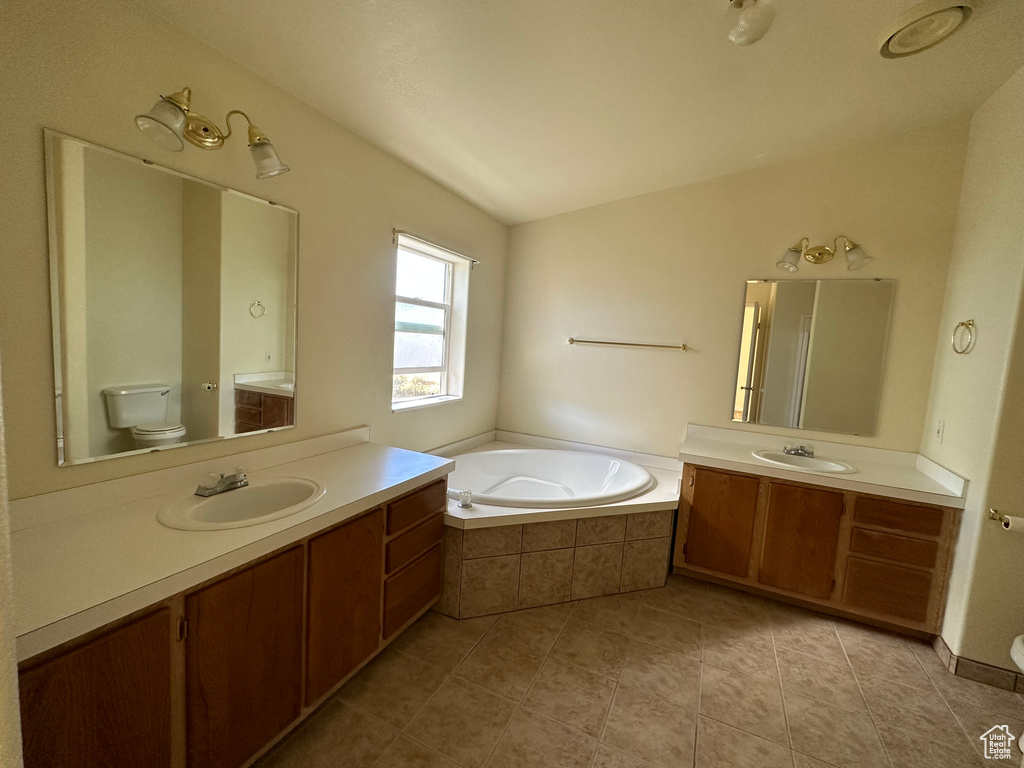 Bathroom with tiled tub, dual bowl vanity, tile floors, and toilet