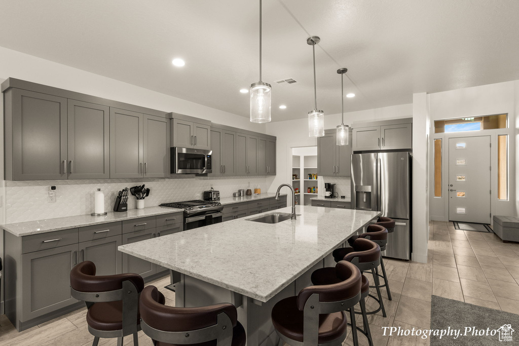 Kitchen with light stone counters, backsplash, hanging light fixtures, appliances with stainless steel finishes, and a kitchen island with sink