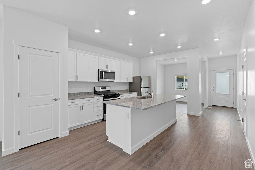 Kitchen with light stone counters, white cabinets, appliances with stainless steel finishes, light hardwood / wood-style floors, and a kitchen island with sink