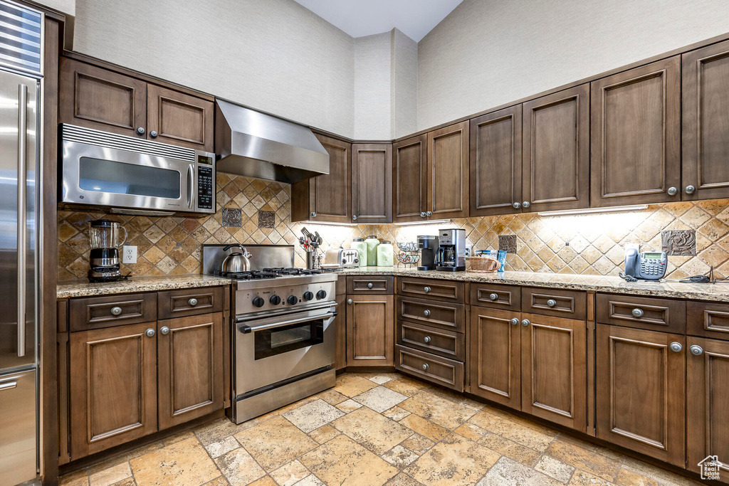Kitchen featuring premium appliances, light stone countertops, backsplash, and wall chimney exhaust hood