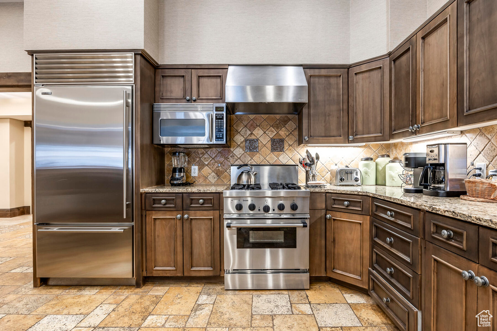 Kitchen with light stone counters, backsplash, light tile floors, high quality appliances, and wall chimney range hood