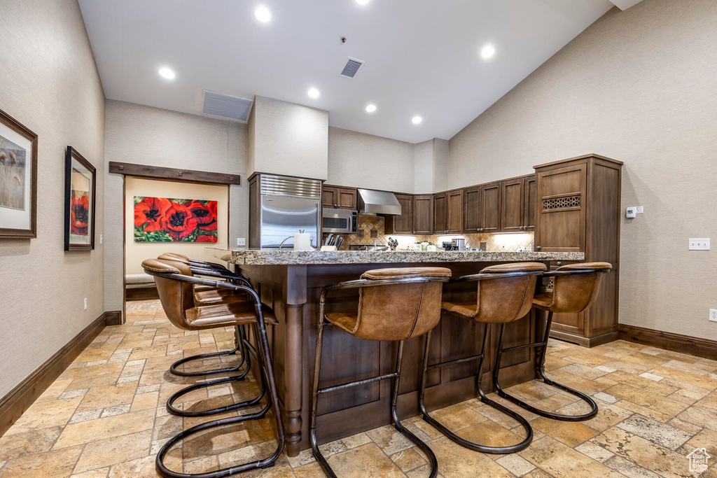 Kitchen featuring wall chimney exhaust hood, high vaulted ceiling, appliances with stainless steel finishes, a kitchen breakfast bar, and light tile floors