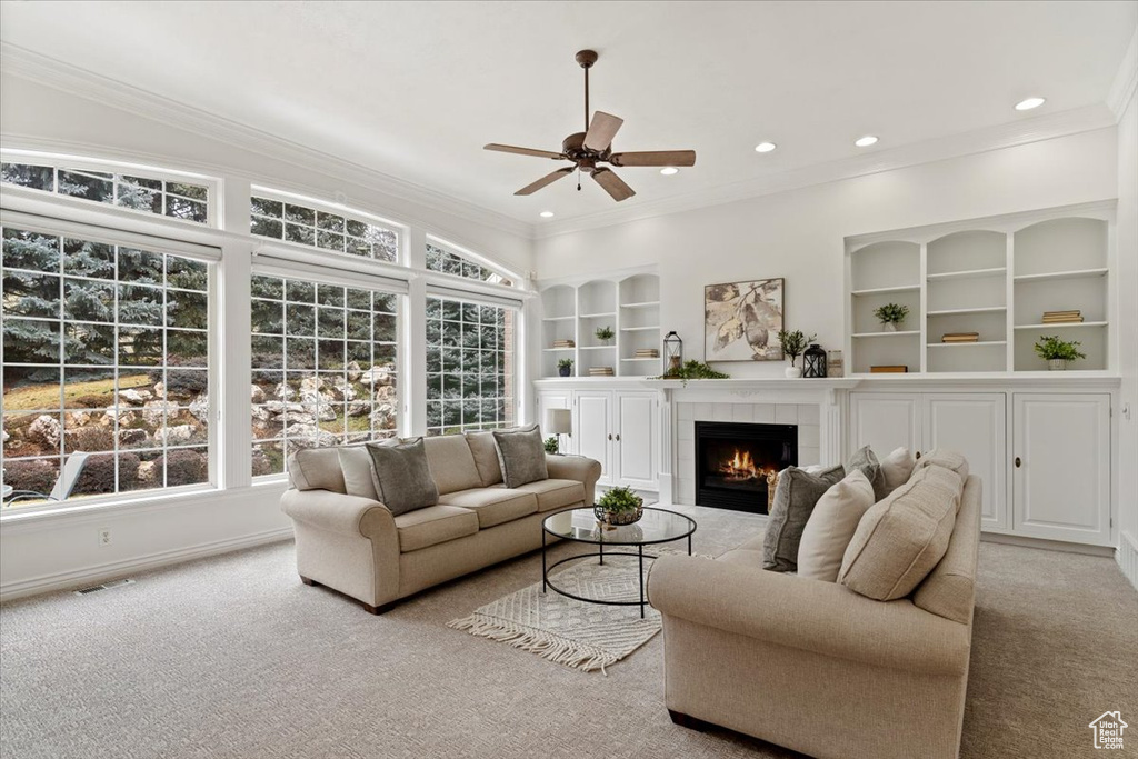 Carpeted living room with crown molding, a tiled fireplace, built in shelves, and ceiling fan
