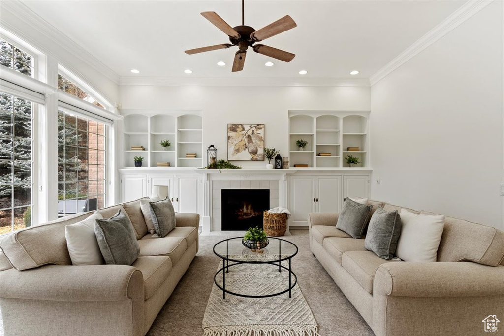 Carpeted living room with a tiled fireplace, built in shelves, ceiling fan, and ornamental molding