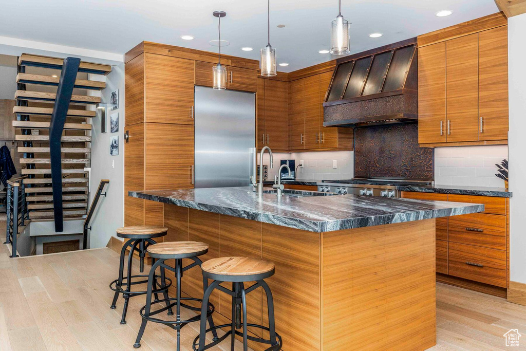 Kitchen featuring premium range hood, light wood-type flooring, tasteful backsplash, stainless steel fridge, and an island with sink