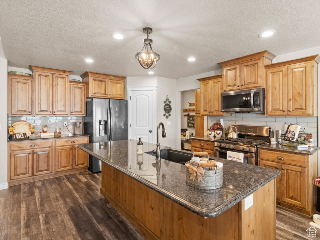 Kitchen with dark wood-type flooring, a center island with sink, appliances with stainless steel finishes, hanging light fixtures, and sink