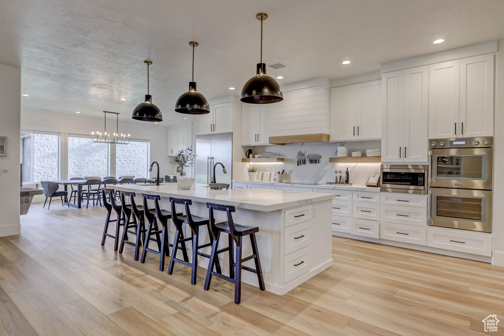 Kitchen with a notable chandelier, white cabinets, a center island with sink, appliances with stainless steel finishes, and light hardwood / wood-style floors