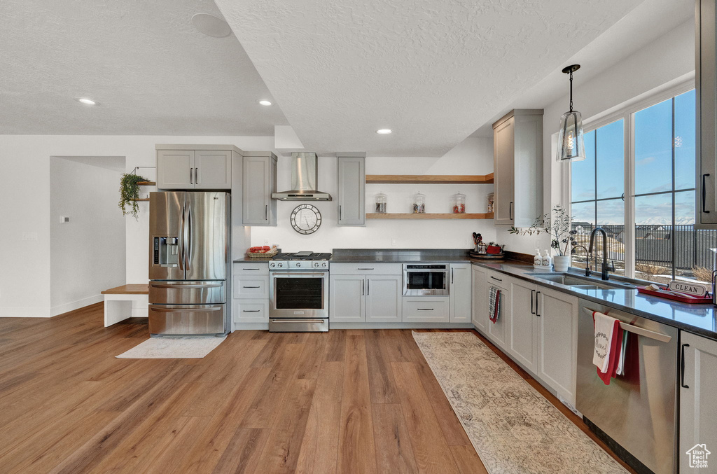 Kitchen with pendant lighting, light wood-type flooring, stainless steel appliances, wall chimney range hood, and sink