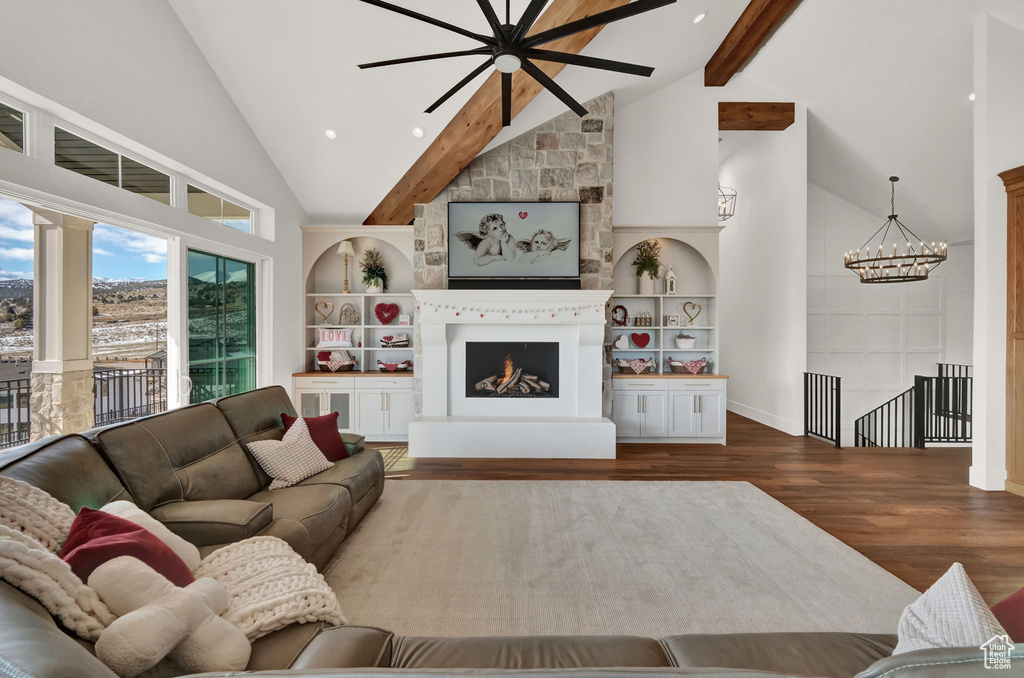 Living room featuring dark wood-type flooring, ceiling fan with notable chandelier, high vaulted ceiling, a stone fireplace, and beam ceiling