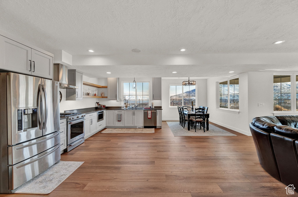 Kitchen featuring light hardwood / wood-style floors, a textured ceiling, hanging light fixtures, stainless steel appliances, and wall chimney exhaust hood