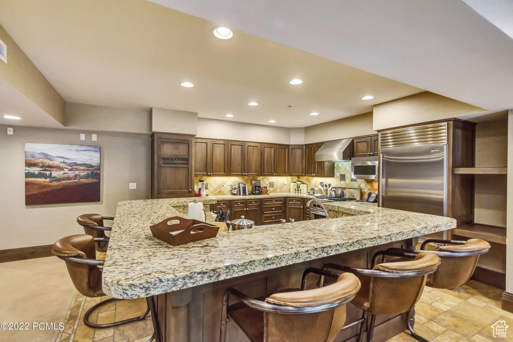 Kitchen featuring tasteful backsplash, a breakfast bar, stainless steel appliances, light tile flooring, and wall chimney range hood