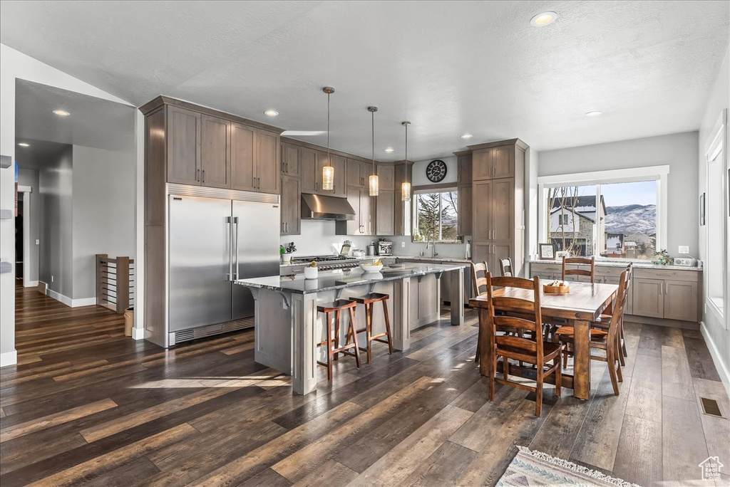 Kitchen featuring dark wood-type flooring, a kitchen island, stainless steel built in refrigerator, hanging light fixtures, and a kitchen breakfast bar